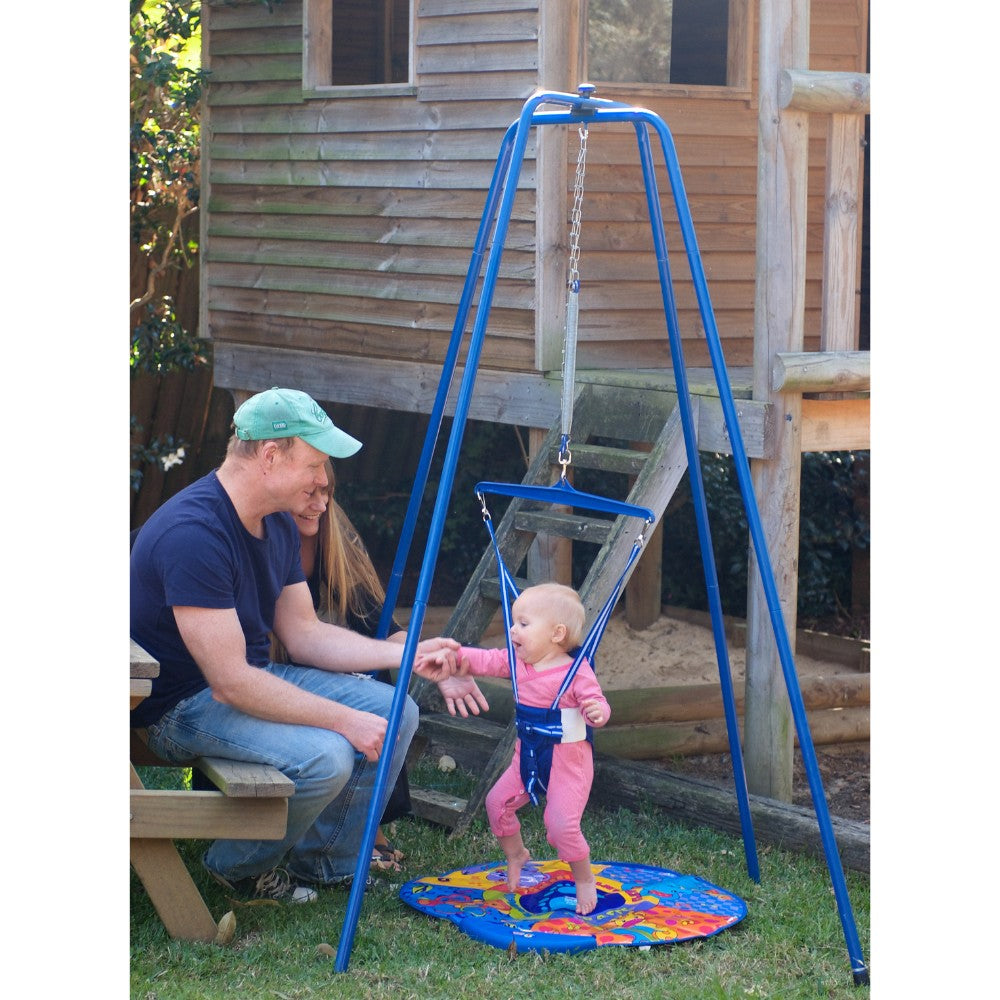 Baby enjoying the Jolly Jumper with Stand in the outdoors, supported by parents near a wooden cubby house
